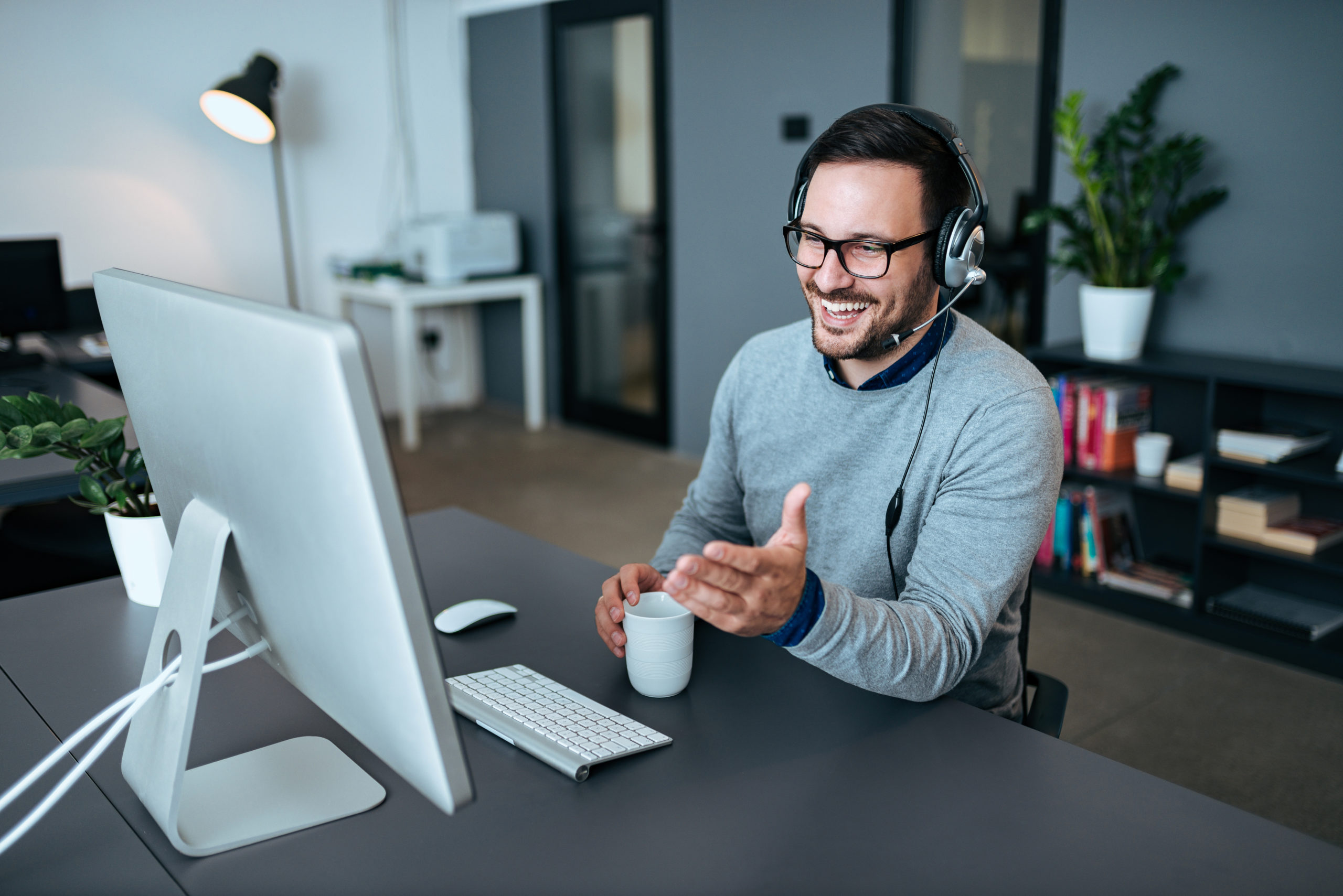 A male sales person sits in front of a large, Apple computer. He is wearing a black headset and smiling as he holds a white cup of coffee.